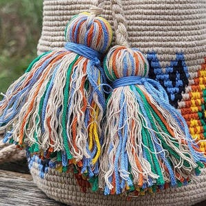 May include: A beige woven bucket bag featuring two large decorative tassels made of multi-colored threads in shades of blue, orange, green, and cream. The bag body displays a geometric pattern in black, yellow, and orange against a beige base.