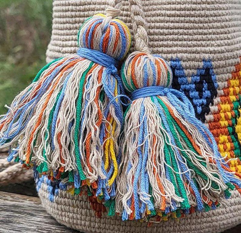 May include: A beige woven bucket bag featuring two large decorative tassels made of multi-colored threads in shades of blue, orange, green, and cream. The bag body displays a geometric pattern in black, yellow, and orange against a beige base.