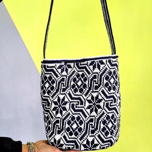 May include: A handmade, cylindrical shoulder bag with a geometric pattern in navy blue and white. The bag has a navy blue strap and trim. The background is a combination of yellow and gray.