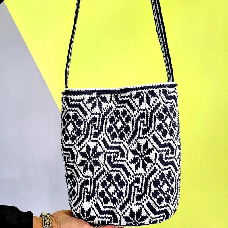 May include: A handmade, cylindrical shoulder bag with a geometric pattern in navy blue and white. The bag has a navy blue strap and trim. The background is a combination of yellow and gray.