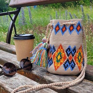 May include: A beige crochet bucket bag featuring a geometric pattern in blue, orange, and yellow, with a matching braided shoulder strap and a multi-colored tassel. Beside it sits a brown paper coffee cup with a black lid and round sunglasses.