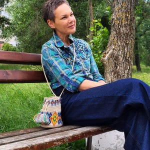 May include: A blue and green plaid long-sleeve button-down shirt with dark blue denim pants and a beige bucket bag featuring a colorful geometric pattern in orange, yellow, and blue, finished with a multi-colored tassel hanging from the side.