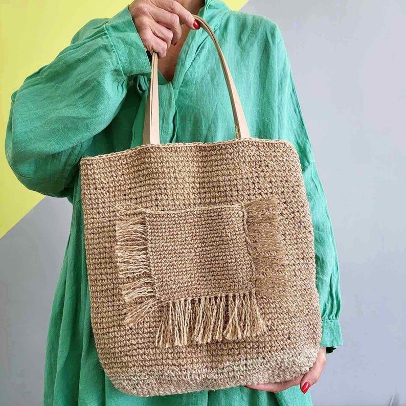 Large handmade natural jute crochet tote bag with fringe front pocket and faux leather shoulder straps.