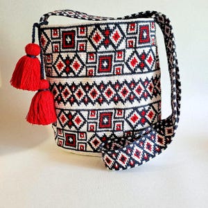 May include: A white, red, and navy blue crocheted shoulder bag with a geometric pattern. The bag has a long strap and two red tassels. The pattern includes diamonds and squares. The bag is approximately 12 inches tall.