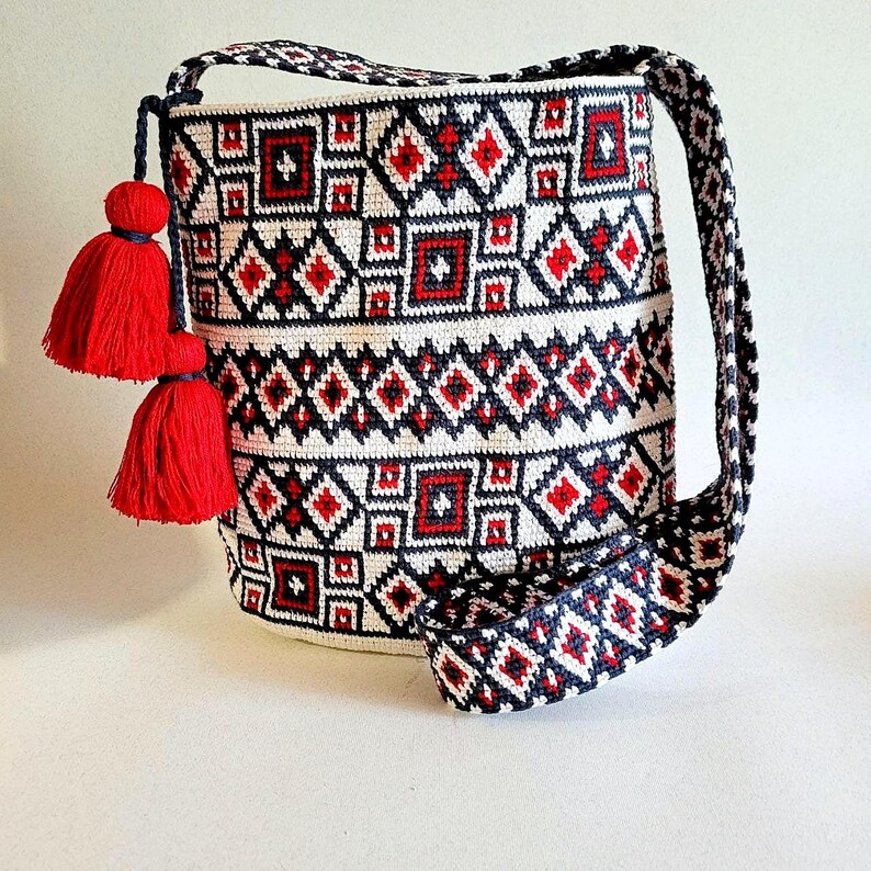 May include: A white, red, and navy blue crocheted shoulder bag with a geometric pattern. The bag has a long strap and two red tassels. The pattern includes diamonds and squares. The bag is approximately 12 inches tall.