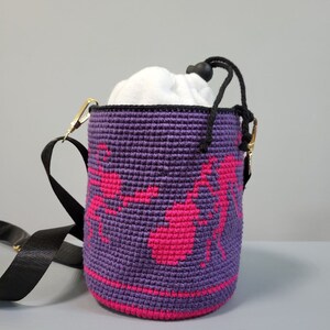 May include: Purple crocheted bucket bag with a pink ant design and a black adjustable strap.