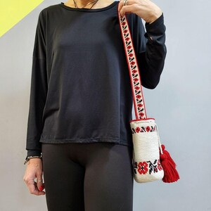 May include: A white crocheted bag with red and black floral embroidery. The bag has a long red and black strap and red tassels.