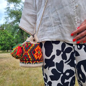 May include: A cylindrical crochet bucket bag featuring a vibrant geometric pattern in red, yellow, and green on a cream base. The bag includes a braided cream shoulder strap and a large, multi-colored tassel made of red, yellow, and green yarn.