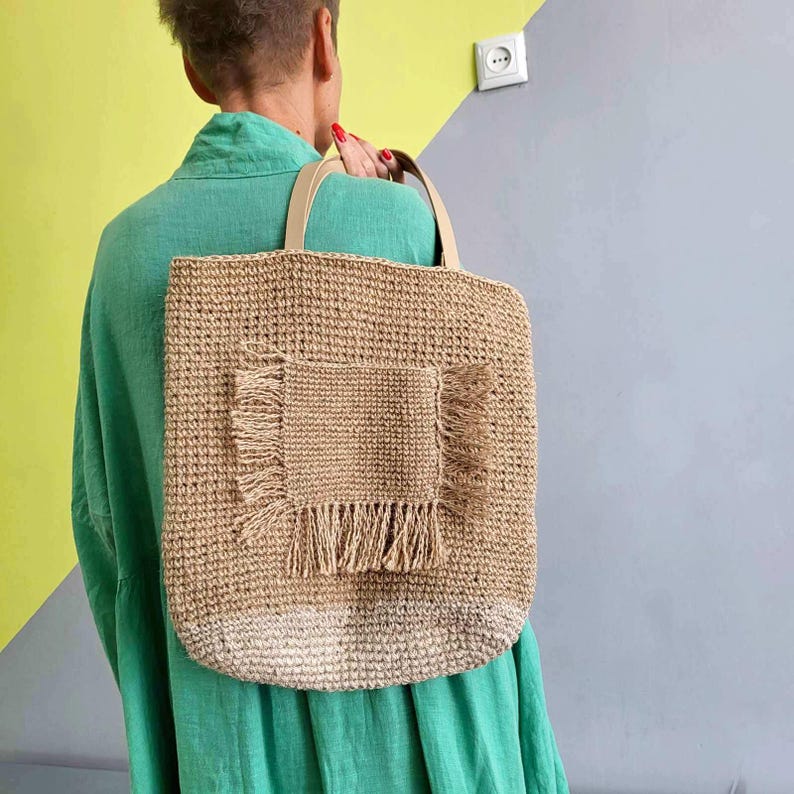 May include: A tan crocheted tote bag featuring a square front pocket with frayed edges and two light-colored handles. The bottom section of the bag has a lighter, off-white color tone compared to the main body, which is a natural tan fiber.