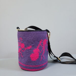May include: A purple crocheted bucket bag with a pink design of ants. The bag has a black adjustable strap.