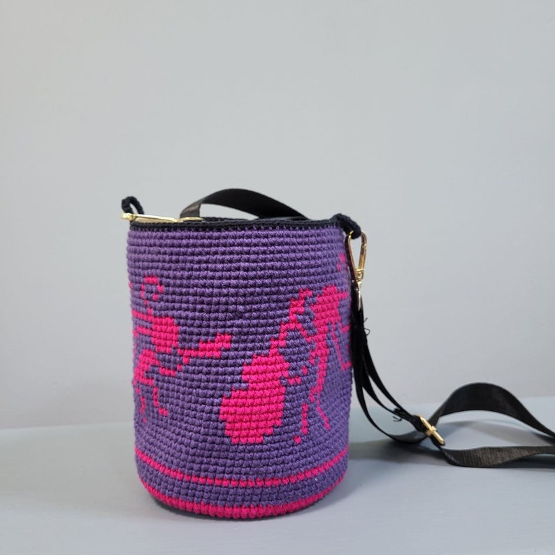 May include: A purple crocheted bucket bag with a pink design of ants. The bag has a black adjustable strap.