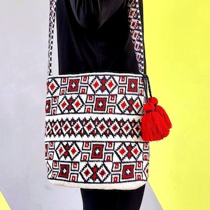 May include: A white, red, and black patterned crossbody bag with a matching strap and red tassels. The bag features a geometric design with diamond and square shapes. The strap and tassels add a decorative touch to this handmade bag.