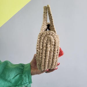 May include: A round, beige, crocheted handbag made of thick, textured natural fiber cord. The bag features two integrated handles at the top and a small, bright red tassel accent attached to the side, showcasing a dense, ribbed stitch pattern.