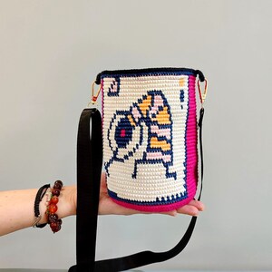May include: A pink and white crocheted bucket bag with a black strap. The bag features a pixelated design of a cartoon character with a yellow and orange striped body.