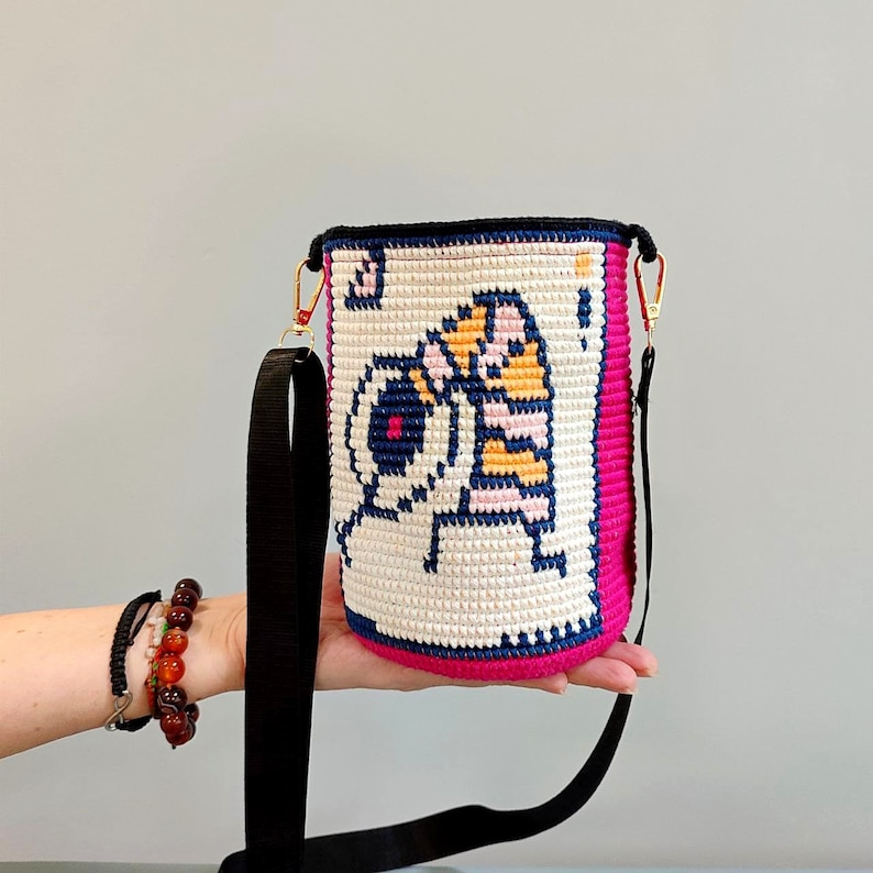 May include: A pink and white crocheted bucket bag with a black strap. The bag features a pixelated design of a cartoon character with a yellow and orange striped body.