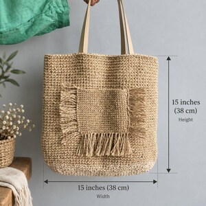 May include: A tan woven tote bag with two light-colored shoulder straps and a central square pocket featuring fringed edges. The bag measures 15 inches in height and 15 inches in width, as indicated by the visible measurement text and arrows.