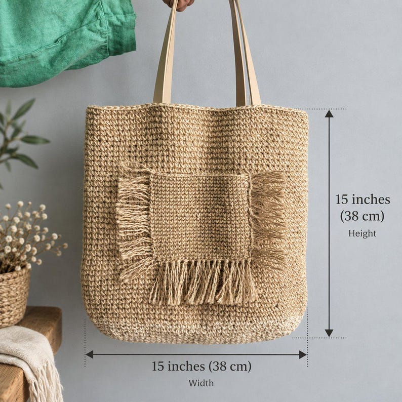 May include: A tan woven tote bag with two light-colored shoulder straps and a central square pocket featuring fringed edges. The bag measures 15 inches in height and 15 inches in width, as indicated by the visible measurement text and arrows.