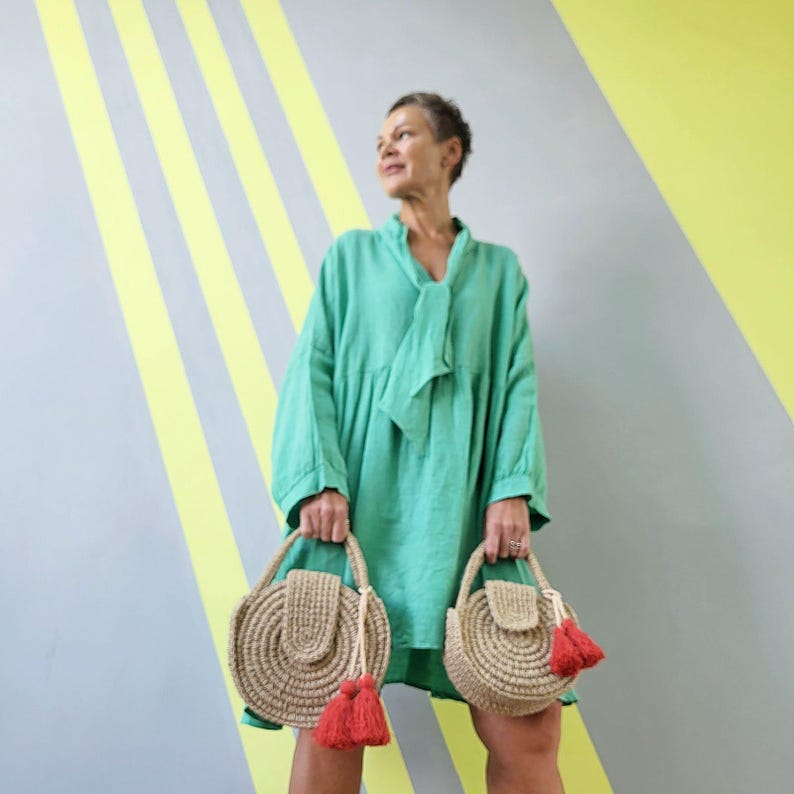 May include: A bright green long-sleeved dress with a tie-neck detail, paired with two round, woven tan straw handbags. Each bag features a structured circular body, a flap closure, and a decorative red tassel attached to the handle.