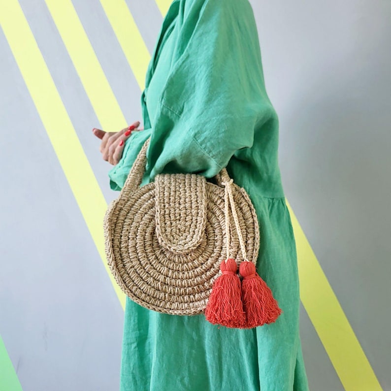 May include: A round, woven straw handbag featuring a textured, circular pattern and a flap closure. The bag is accented with two long, bright red decorative tassels hanging from a braided cord, providing a vibrant contrast to the natural tan straw.
