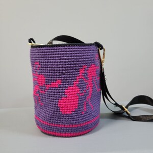May include: A purple crocheted bucket bag with a pink design and a black adjustable strap. The bag has a gold clasp.