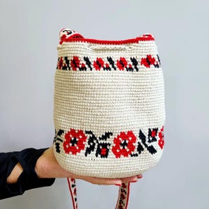 May include: A white crocheted bucket bag with red and black floral embroidery. The bag has a long strap.