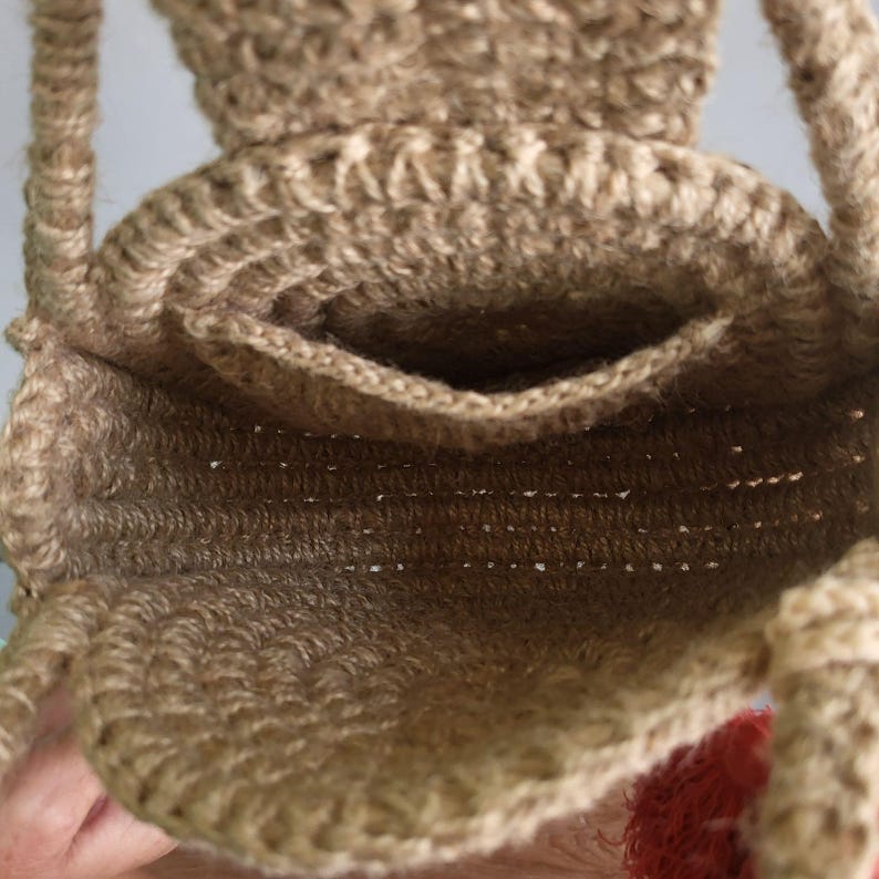 May include: A tan crochet bag made from natural fiber, featuring a circular base, a structured opening with a small interior pocket, and two thick braided handles. The bag has a textured, woven appearance with visible stitching throughout the design.