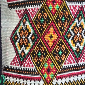 May include: A woven textile featuring a repeating geometric pattern of diamonds and triangles. The design incorporates vibrant colors including black, white, red, orange, yellow, and green threads, creating a dense, textured, and intricate surface.