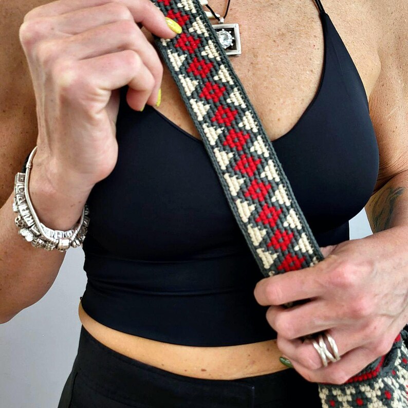 May include: A close-up of a gray and beige woven bag strap with red accents. The strap is being held by a person wearing a black top.