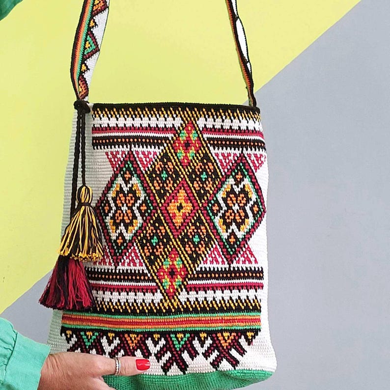 May include: A woven shoulder bag featuring a cream-colored base with intricate geometric patterns in black, orange, pink, and green. The bag includes a long matching strap and a decorative tassel with black, yellow, and red threads hanging from the side.