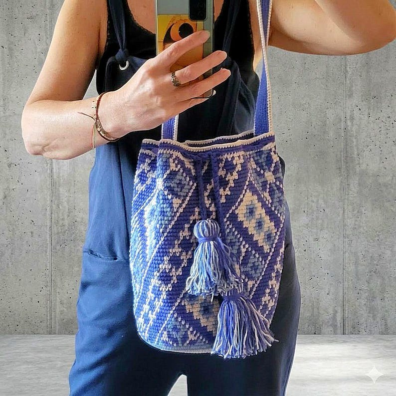 May include: A blue and white crochet shoulder bag with a geometric pattern. The bag has a drawstring closure and two tassels. The strap is blue and white. The bag is held by a person.
