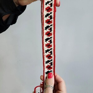 May include: A close-up of a red, white and black crocheted trim. The trim has a geometric pattern and is made with thick yarn.