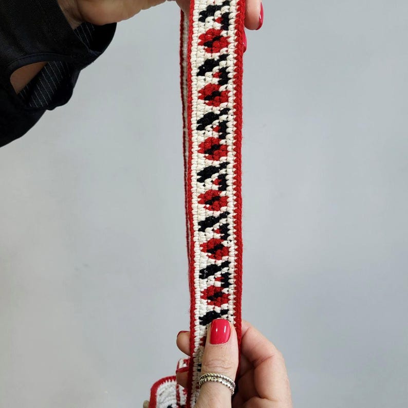 May include: A close-up of a red, white and black crocheted trim. The trim has a geometric pattern and is made with thick yarn.