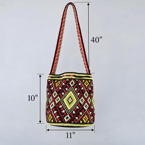 May include: A crochet bucket bag featuring a geometric diamond pattern in black, red, yellow, and white. The bag has a long shoulder strap and measures 10 inches in height, 11 inches in width, and has a total strap length of 40 inches.
