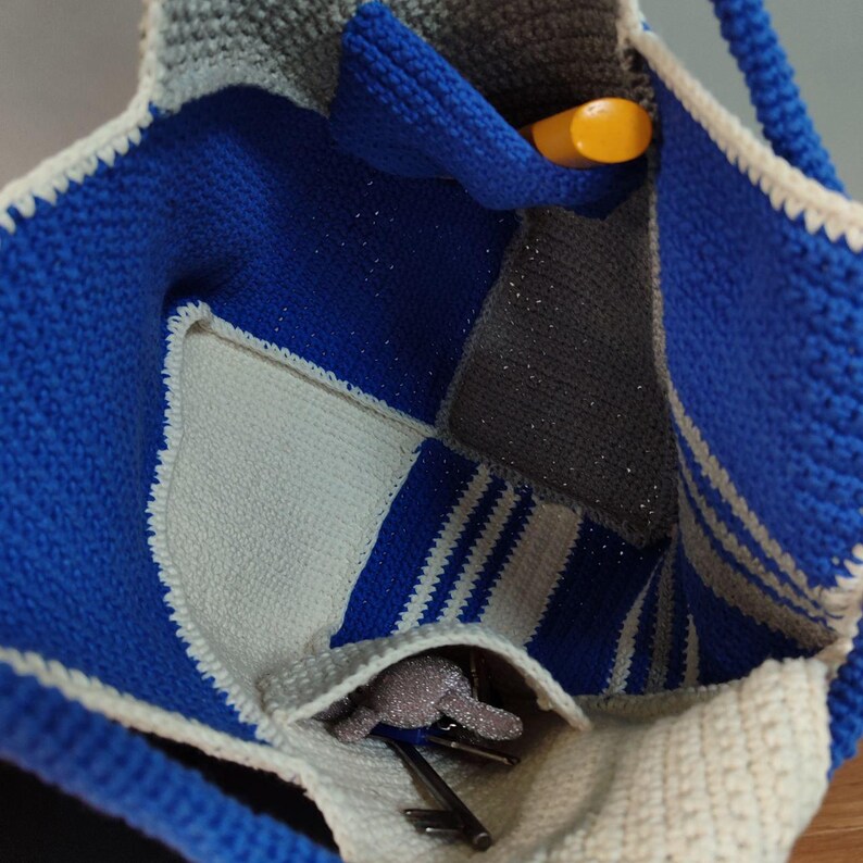 May include: A crocheted tote bag with a patchwork design in blue, white, and gray. The bag has a single handle and a zippered pocket on the inside.