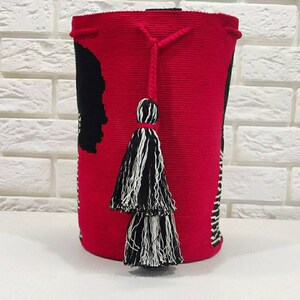 May include: A red crocheted bag with black and white stripes and a black silhouette design. The bag has two black and white tassels.