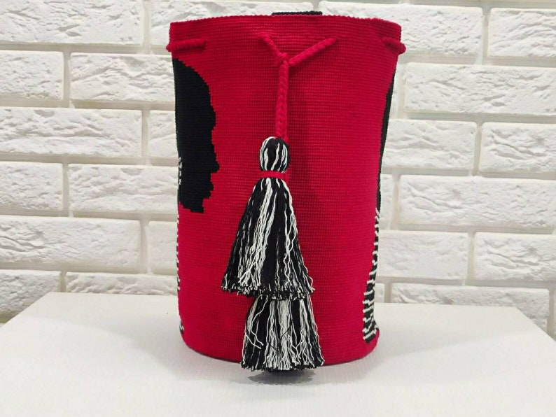 May include: A red crocheted bag with black and white stripes and a black silhouette design. The bag has two black and white tassels.