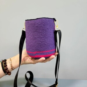 May include: A small purple crocheted bag with a pink stripe and a black adjustable strap. The bag is being held in a hand.