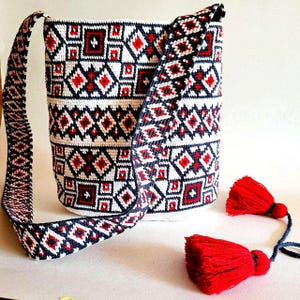 May include: A white, red, and navy blue crocheted shoulder bag with a geometric pattern. The bag has a matching strap and two red tassels. The bag's design features diamond and square shapes.