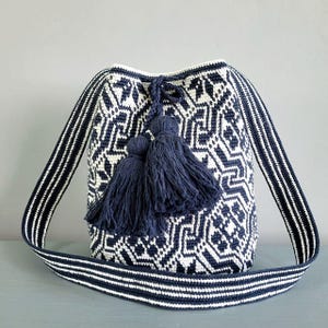 May include: A navy blue and white patterned shoulder bag with a geometric design. The bag features a long strap with white stripes and navy blue tassels. The bag is made of woven material.