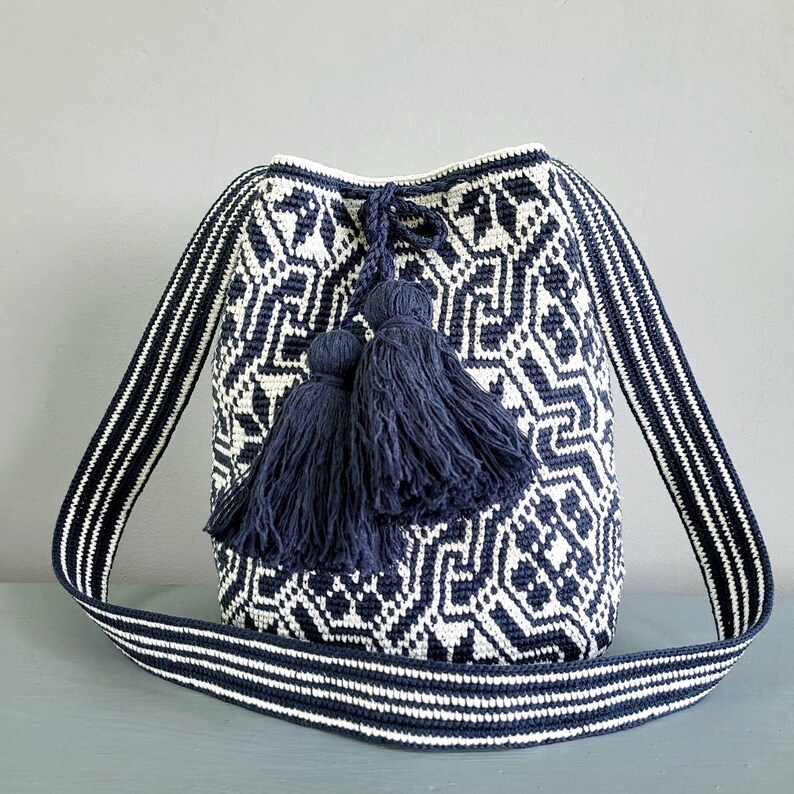 May include: A navy blue and white patterned shoulder bag with a geometric design. The bag features a long strap with white stripes and navy blue tassels. The bag is made of woven material.