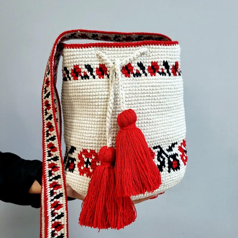 May include: White crocheted bucket bag with red and black embroidered floral design. The bag has a long strap and red tassels.