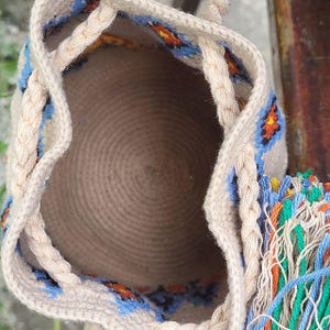 May include: A beige woven bucket bag featuring a circular base and a drawstring closure made of braided cord. The exterior displays a repeating geometric pattern in blue, orange, and black, complemented by a multi-colored tassel attachment.