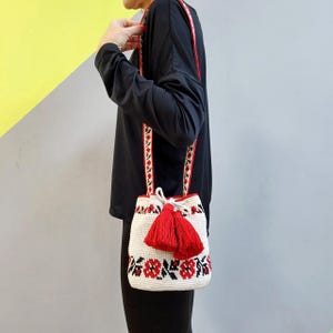 May include: A white crocheted bucket bag with red floral embroidery and red tassels. The bag has a long strap and is worn over the shoulder.