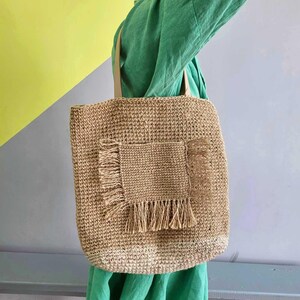 May include: A tan woven tote bag featuring a rectangular front pocket with frayed edges and fringe detailing along the sides and bottom. The bag has two matching tan shoulder straps and a textured, natural fiber construction throughout the body.