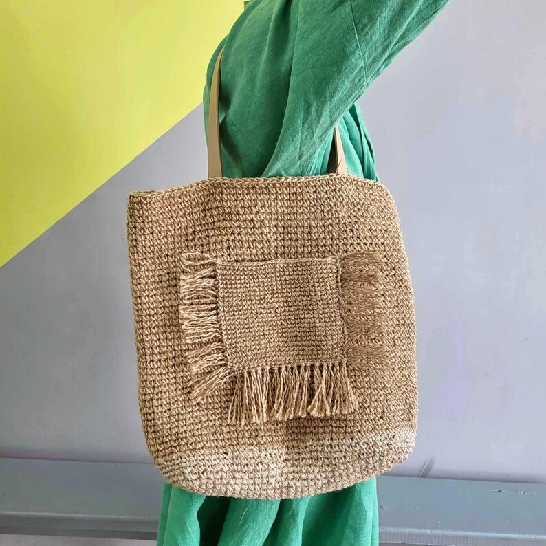May include: A tan woven tote bag featuring a rectangular front pocket with frayed edges and fringe detailing along the sides and bottom. The bag has two matching tan shoulder straps and a textured, natural fiber construction throughout the body.