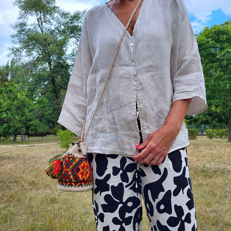 May include: A beige linen button-down shirt with three-quarter sleeves, paired with black and white floral print trousers. A small, cylindrical woven crossbody bag features a vibrant geometric pattern in red, green, and yellow with a tassel.