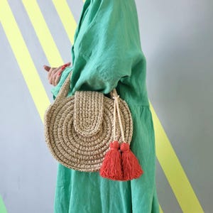 May include: A round, beige, crocheted straw handbag featuring a structured flap closure and two prominent, vibrant orange-red tassels hanging from a braided cord. The bag has a circular woven texture and is carried by a matching crocheted handle.