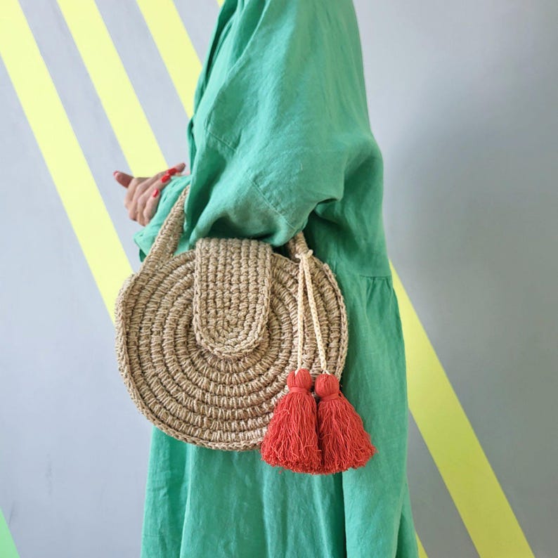 May include: A round, beige, crocheted straw handbag featuring a structured flap closure and two prominent, vibrant orange-red tassels hanging from a braided cord. The bag has a circular woven texture and is carried by a matching crocheted handle.