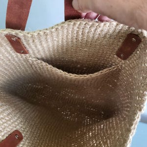 May include: A beige woven jute tote bag featuring brown leather handles attached with silver-toned metal rivets. The interior of the bag is visible, showcasing a textured, natural fiber construction with a single open pocket on the inner wall.