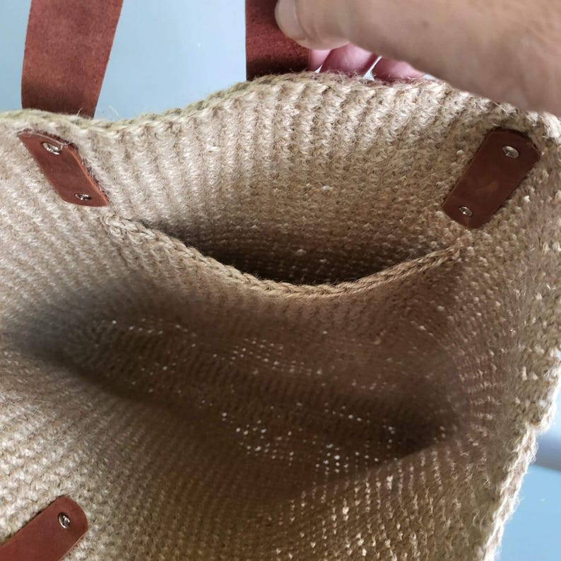 May include: A beige woven jute tote bag featuring brown leather handles attached with silver-toned metal rivets. The interior of the bag is visible, showcasing a textured, natural fiber construction with a single open pocket on the inner wall.